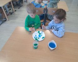 Przedszkolaki tworzą kreatywne prace plastyczne przy stolikach, a następnie sadzą drzewa w ogrodzie.